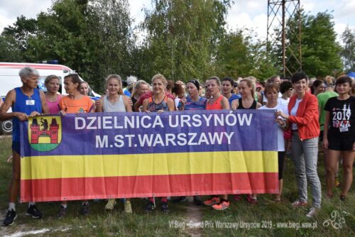 Grupa kobiet i mężczyzna trzymają flagę dzielnicy Ursynów w Warszawie.